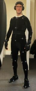 Performance Artist, Scott Joseph in a blue figure hugging motion capture suit that is covered with reflective balls. He is standing in an overly dramatic pose and is wearing motion capture gloves. A small microphone is positioned high on his forehead and he is wearing over the ear headphones.