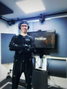 Performance Artist, Scott Joseph in a black figure-hugging motion capture suit that is covered with reflective balls. The photos is cropped to three-quarter length and he is standing with his arms folded and smiling. He is wearing motion capture gloves. A small microphone is positioned high on his forehead attached to a material skull cap and he is wearing over-the-ear headphones. He is posing next a monitor that has Pitstop Productions on the screen and the walls of the room are acoustically treated and have motion cameras dotted about.