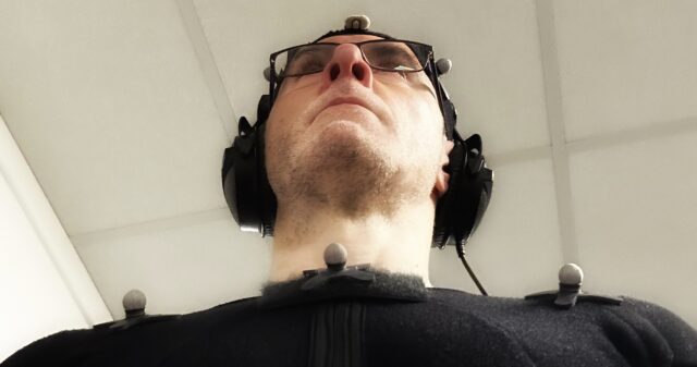 A stylised mid shot of Voice and Performance Artist Scott Joseph from a low angle with him wearing a black motion capture suit and headphones and staring past the top of the camera.