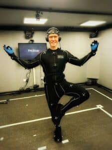 Performance Artist, Scott Joseph in a black figure-hugging motion capture suit that is covered with reflective balls. The photos is cropped to three-quarter length and he is standing on one leg in a mock meditation pose, the other leg bent out to the side, foot resting on the standing leg's knee. His arms are held out wide with the index fingers on each hand touching their respective thumbs. He's smilng. He is wearing motion capture gloves. A small microphone is positioned high on his forehead attached to a material skull cap and he is wearing over-the-ear headphones. He is posing next a monitor that has Pitstop Productions on the screen and the walls of the room are acoustically treated and have motion cameras dotted about.